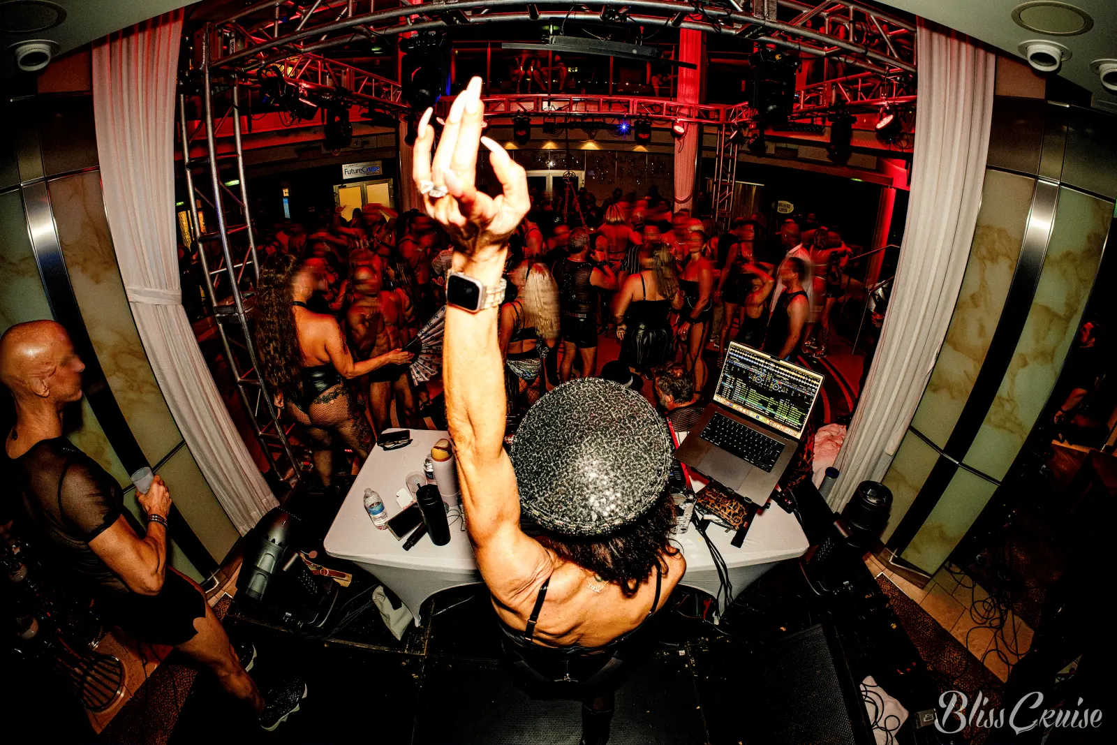 A DJ with an arm raised, seen from behind, facing a dancing crowd in the ship nightclub