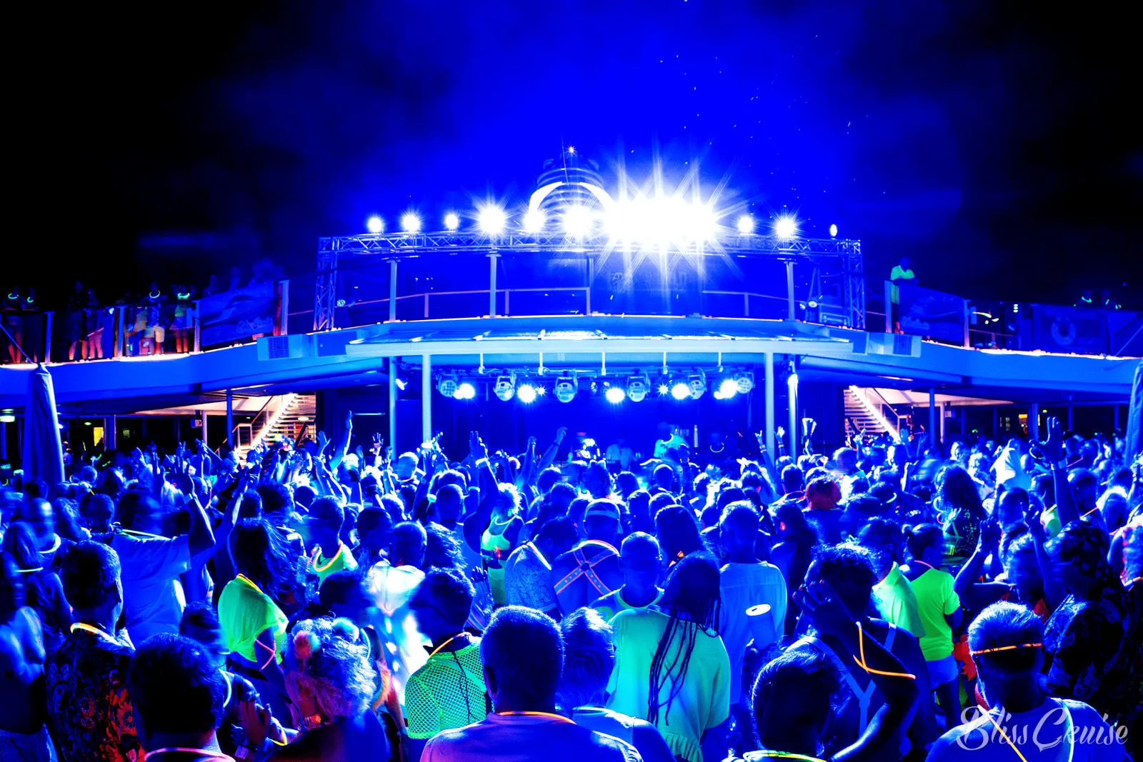 A crowd in neon glow accessories dancing at a night party on the pool deck under blue stage lights